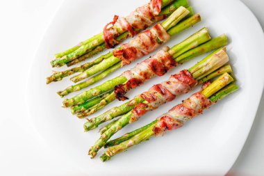 Grilled green asparagus wrapped with bacon. Asparagus baked with bacon and spices. Healthy food. Top view. Free space for your text. Bacon wrapped asparagus on white plate.
