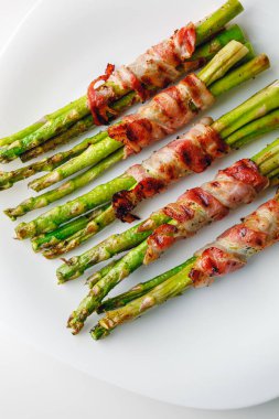 Grilled green asparagus wrapped with bacon. Asparagus baked with bacon and spices. Healthy food. Top view. Free space for your text. Bacon wrapped asparagus on white plate.