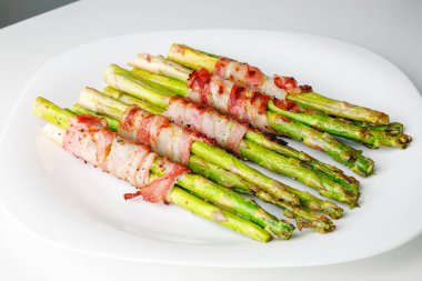 Grilled green asparagus wrapped with bacon. Asparagus baked with bacon and spices. Healthy food. Top view. Free space for your text. Bacon wrapped asparagus on white plate.