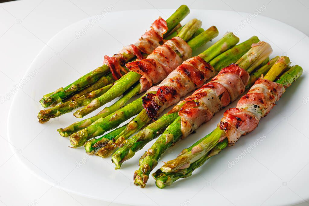 Grilled green asparagus wrapped with bacon. Asparagus baked with bacon and spices. Healthy food. Top view. Free space for your text. Bacon wrapped asparagus on white plate.