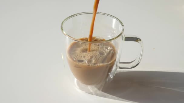 Verter café fresco a la taza de vidrio transparente con hielo en la mesa blanca, gotas — Vídeo de stock