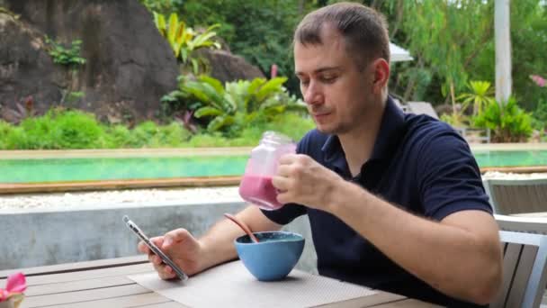 Junger positiver Mann hält Smartphone, liest Nachrichten online aufmerksam, trinkt Smoothie — Stockvideo