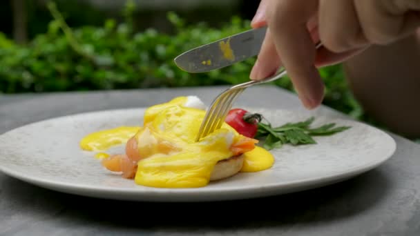 Close-up man hands with fork and knife cuts eggs benedict in outdoor restaurant — Stock Video