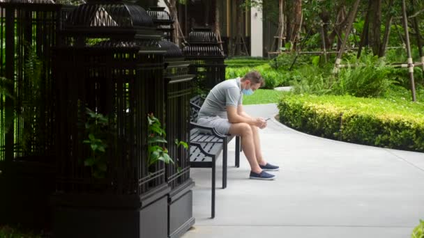 Młody dorosły mężczyzna nosi maskę, siedzi skulony w parku, pracuje ze smartfonem — Wideo stockowe