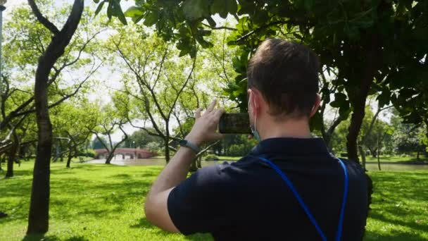 Vista trasera del hombre influencer en el parque fotografiando con lago y puente — Vídeos de Stock