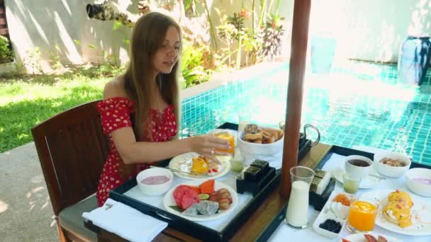 Nuori nainen syö aamiaista uima-altaan lähellä, jossa on sininen vesi huvilalla — kuvapankkivideo