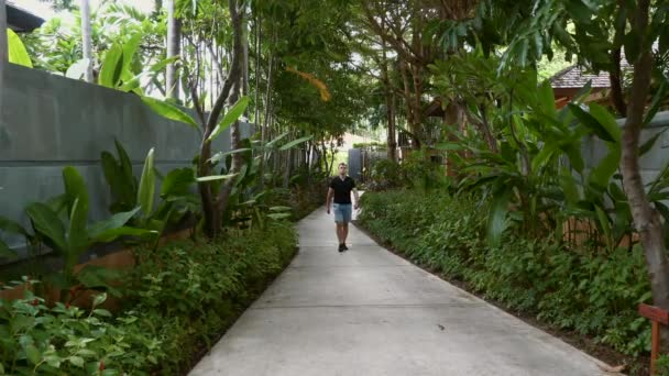 Travel man in casual walking on path among palm trees in modern luxury resort — Stock Video