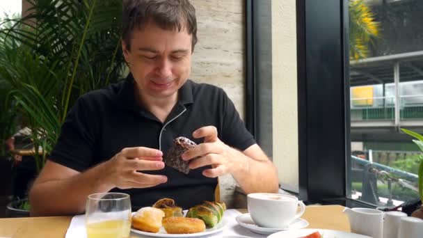 Jovem com fome mordendo muffin, mastigar e desfrutar do sabor, sente-se no café — Vídeo de Stock