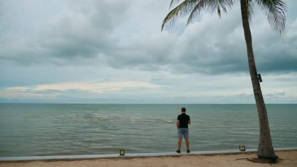 Pora deszczowa w Tajlandii. Podróżnik stojący na plaży z palmą — Wideo stockowe