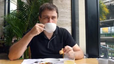 Gülümseyen adam kahve içiyor ve şehirdeki kafede taze fırınla kahvaltı ediyor.