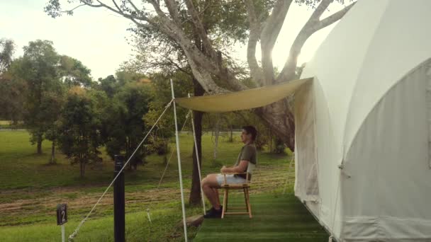 Joven turista viajero hombre disfrutando de la naturaleza, sentado cerca de lona tienda de campaña moderna — Vídeo de stock