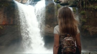 Female backpacker tourist looking at waterfall in jungles, touches her long hair