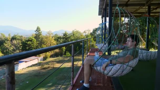 Hombre salones y relajarse en la silla colgante en la cafetería con vista a las montañas — Vídeo de stock