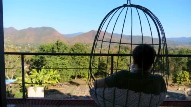 Back view of male tourist sitting on hanging chair in cafe, drinking coffee