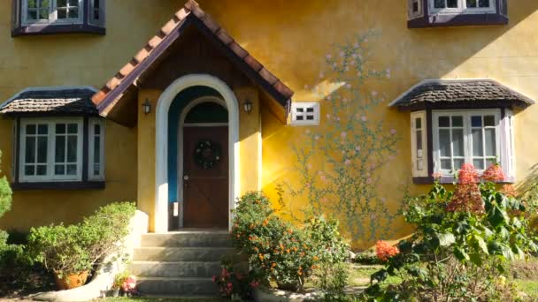 Желтый рустистый дом с цветущими цветами, зелеными листьями и красивыми окнами — стоковое видео