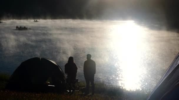 Пара мандрівників, що стоять біля намету в кемпінгу, розмовляють біля туманної річки — стокове відео