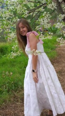 Beyaz sakura çiçekleri arasında yürüyen güzel ve genç bir kadının videosu. Gülümseyen kadın kiraz ağaçlarının çiçeklerini sever. Parkta sakura ağacının bahar çiçeği. Güzellik kavramı, kozmetik. 
