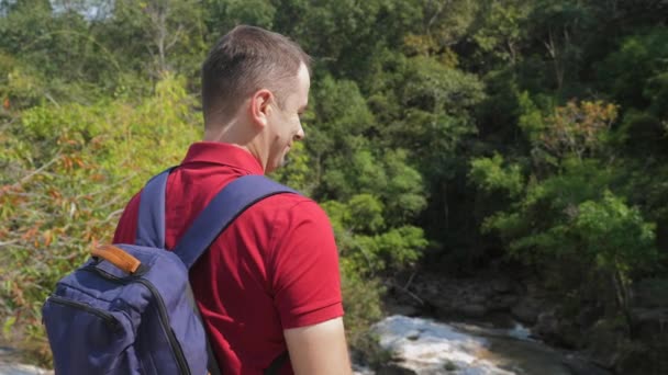 旅行男で赤いポロシャツにリュックを岩の川の近くに立っている — ストック動画
