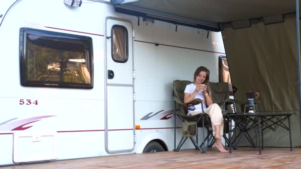 Une jeune femme souriante est assise sur une chaise de camping pliante près d'une caravane — Video