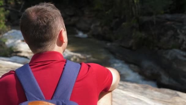 Primer plano del viajero masculino con mochila en su espalda sentado cerca del río de montaña — Vídeo de stock