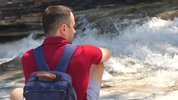 Alone male traveler with backpack sits near bubbling mountain river — Stock Video