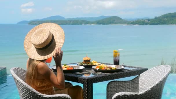 Γυναίκα σε ψάθινο καπέλο κοντά στην πισίνα, τρώγοντας φαγητό και απολαμβάνοντας θέα στον ωκεανό — Αρχείο Βίντεο