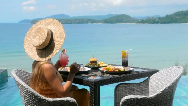 Путешествующая женщина в соломенной шляпе пьет коктейль на обед с видом на море — стоковое видео