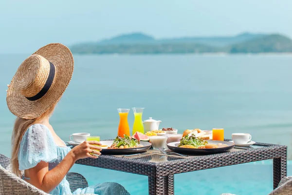 Yüzme havuzunun yanındaki hasır şapkalı kadın yemek yiyor ve okyanus manzarasının tadını çıkarıyor. — Stok fotoğraf