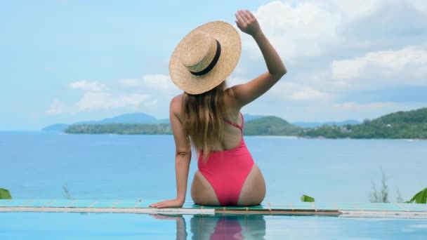 Σέξι γυναίκα με μαγιό και ψάθινο καπέλο που κάθεται στην άκρη της πισίνας — Αρχείο Βίντεο