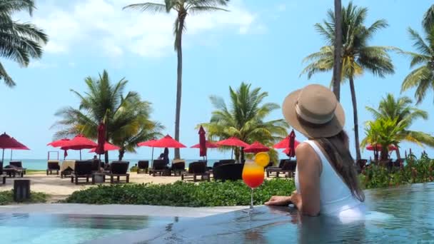 Reisefrau in Hut und Badebekleidung mit Erfrischungsgetränk schwimmt im Pool — Stockvideo