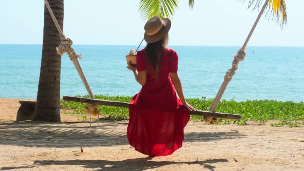 Jonge alleenstaande vrouw rust op schommel op het strand, houd vers geopende kokosnoot in de hand — Stockvideo