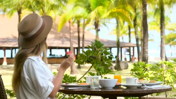 Молодая женщина в шляпе ест свежий арбуз, завтракает в курортном ресторане — стоковое видео