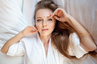 Yatakta beyaz bornozlu genç bir kadın oteldeki ya da otel odasındaki kameralara bakıyor.