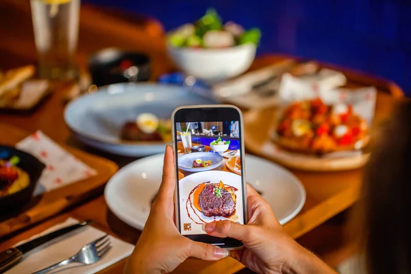 Lüks restoran masasında cep telefonuyla gurme bifteği çeken bir kadın.