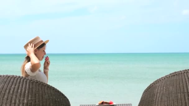 Ελκυστική νεαρή γυναίκα με ψάθινο καπέλο κάθεται στην παραλία καφέ στη θάλασσα, τρώνε καρπούζι — Αρχείο Βίντεο