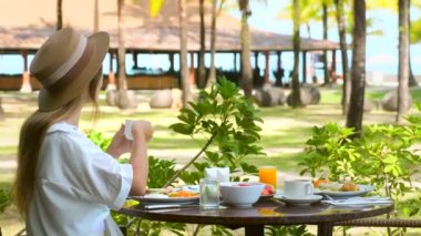 Genç bir kadın açık havada lüks bir restoranda beyaz bardakta sıcak kahve içiyor.