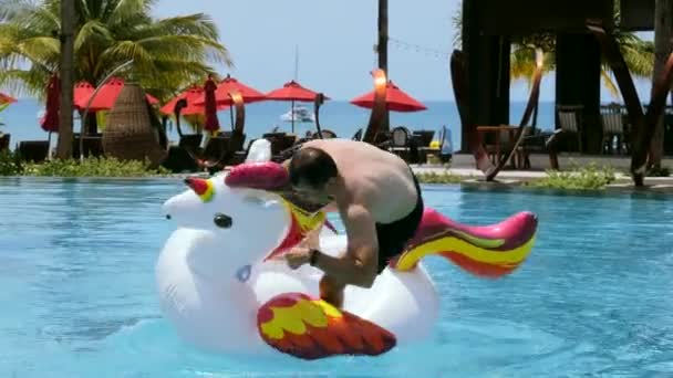 Happy man have fun ride on giant inflatable unicorn, splashing in swimming pool — стоковое видео