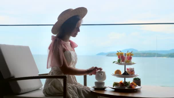 Κομψή κυρία σε λευκό φόρεμα και ψάθινο καπέλο ρίχνει ζεστό τσάι και να απολαύσετε γλυκό φαγητό — Αρχείο Βίντεο