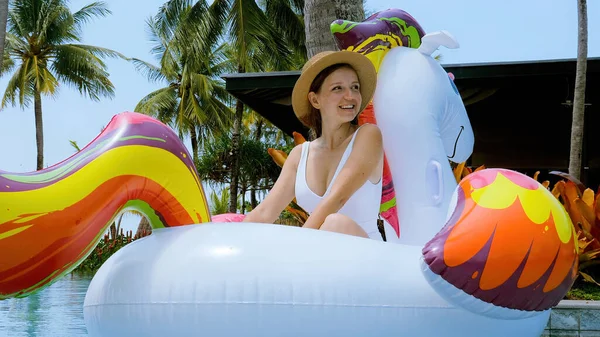 Happy traveler tourist in swimwear and hat relaxing on inflatable unicorn toy at holiday at luxury hotel tropical resort swimming pool. Enjoying tropical summer, travel in Thailand, tourism lifestyle. — Zdjęcie stockowe