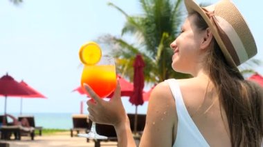 Beautiful girl in swimwear drinking colorful yummy cocktail at swimming pool. Woman in white swimsuit drinking non-alcoholic orange cocktail, relax in pool water, enjoy vacation at luxury hotel.