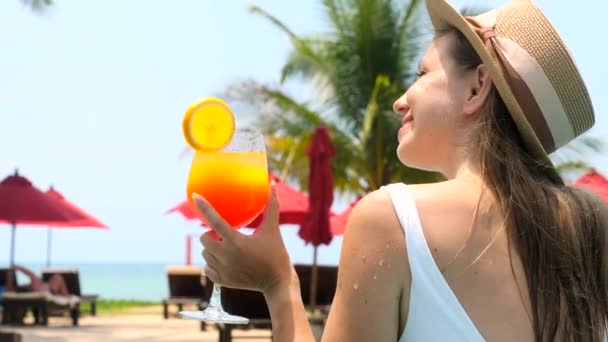Beautiful girl in swimwear drinking colorful yummy cocktail at swimming pool. Woman in white swimsuit drinking non-alcoholic orange cocktail, relax in pool water, enjoy vacation at luxury hotel. — Stock videók
