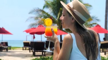 Havuzda güneşli bir günde ferahlatıcı bir yaz kokteyli içen genç ve güzel bir kadın. Mutlu gülümseyen bayan gezgin deniz manzaralı tropikal bir tatil köyünde dinleniyor. Plaj kenarı oteli.