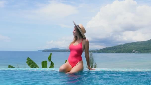 Travel girl in straw hat and swimsuit sitting by pool side at sunny summer day — Stockvideo