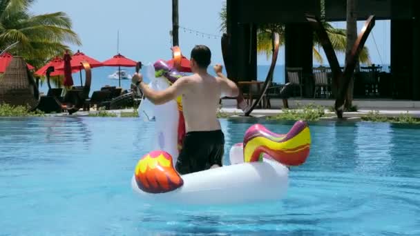 Funny video young man falling in pool in luxury hotel, resort. Happy man have fun ride dance on inflatable unicorn in swimming pool, splashing, enjoy holidays or vacations on sunny day in summer — Vídeo de Stock