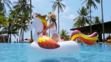 Tayland 'da seyahat. Mutlu, gülümseyen kadın eğlencesi, yüzme havuzunda şişme tek boynuzlu at yatağında dans etmek ve tropikal bir tatil olan palmiye ağaçları ve deniz kıyısında yaz tatilini kutlamak.