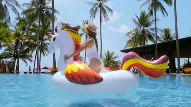 Traveling in Thailand. Happy smiling woman fun, dancing on big inflatable unicorn toy mattress in swimming pool and celebrating summer vacation in tropical resort with palm trees and sea — Stockvideo