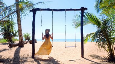Sahilde salıncakta sallanan şapkalı gezgin bir kadın. Palmiye ağaçlarıyla.