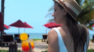 Elinde tropikal kokteyl olan bir kadının arkası. Palmiye ağaçları ve deniz manzaralı kumlu sahilin yakınındaki yüzme havuzunda dinleniyor ve serinliyor. Tatil kavramı.