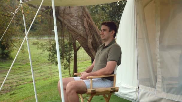 Camp site in forest. Handsome man relax and enjoy nature, sitting near modern canvas tent tourist camp. Male tourist enjoying leisure and freedom. Traveling, vacation, birdwatching, camping concept. — Stock Video