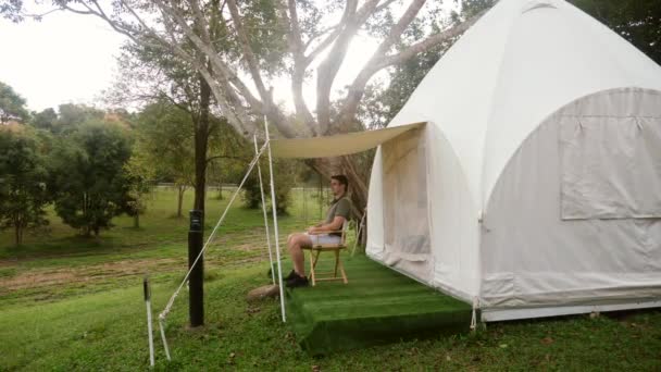Ung turist rejsende mand nyder naturen, sidder på stol nær lærred moderne lejr telt på skov camping. Aktiv livsstil, rejser, turisme, dyreliv ser. Nationalpark, eventyr. – Stock-video